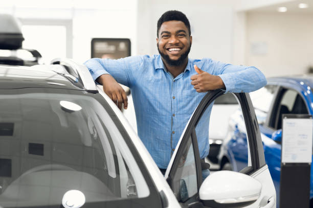 Car Buyer. African Man Gesturing Thumbs Up Approving Automobile After Test Drive Standing In Dealership Center. Selective Focus