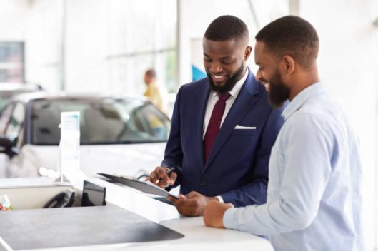 Joyful Black Man Buying Car, Signing Papers With Salesman In Dealership Office, Happy Young African American Male Consulting With Manager While Purchasing New Vehicle In Modern Showroom, Copy Space